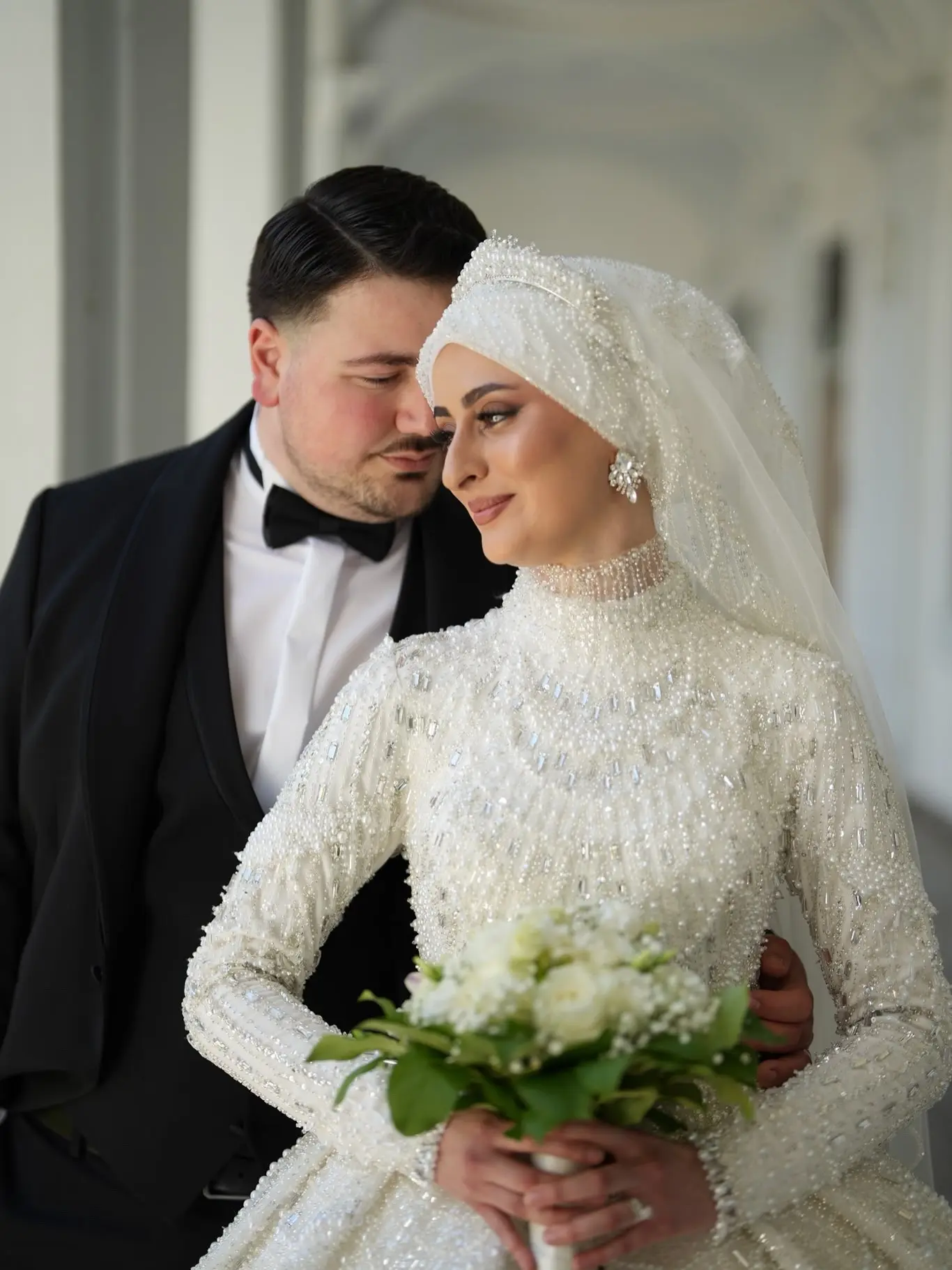 Braut mit perlenbesticktem weißen Hijab und Schleier, dahinter der Bräutigam